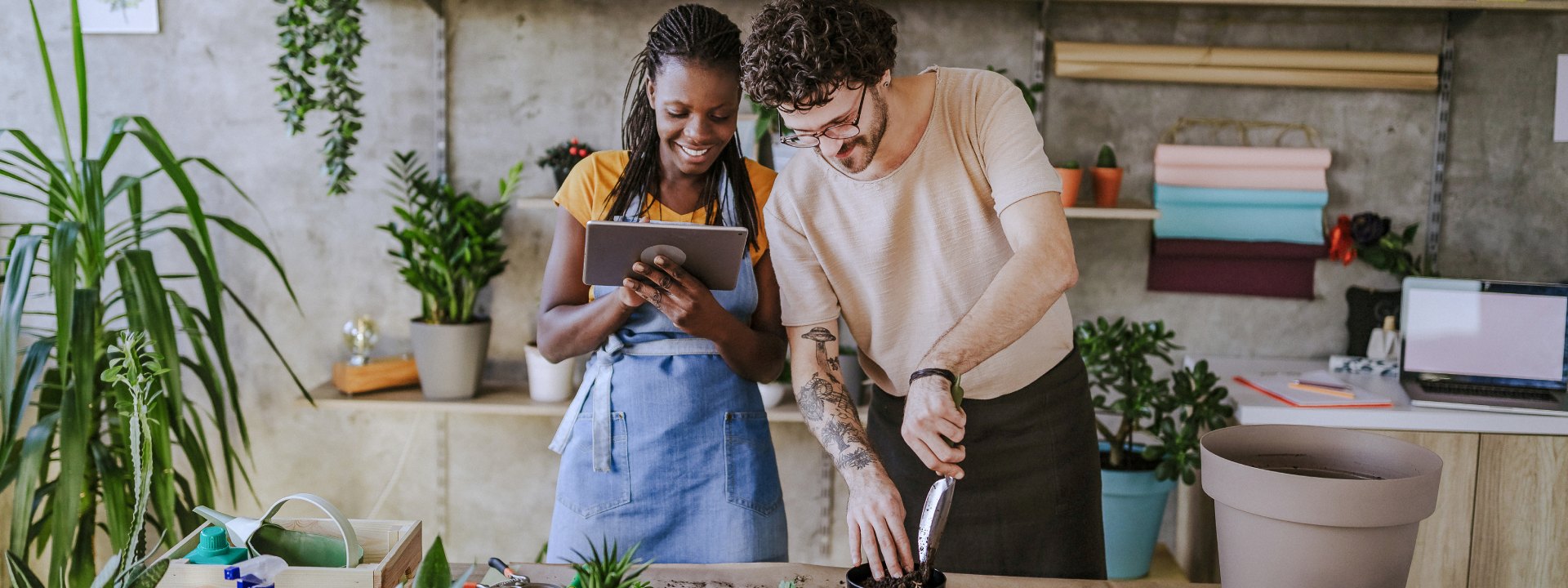 Two people potting plants together at a table in a room with shelves of potted plants and gardening tools.
