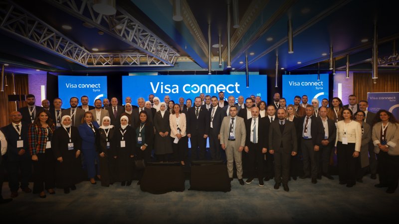 A large, diverse group of professionals poses together at a conference. They stand in front of blue screens displaying "Visa Connect Syria." 