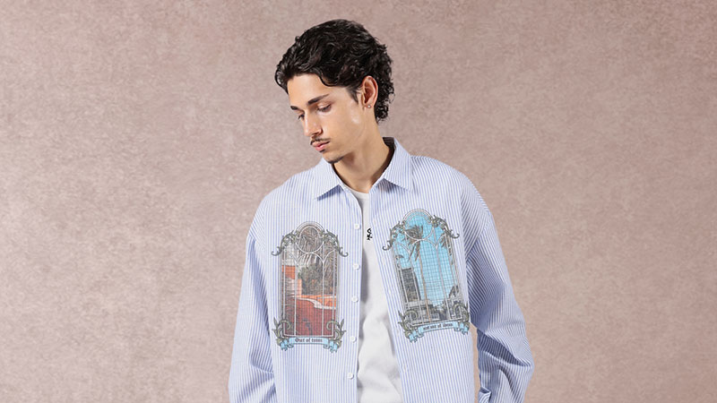 A person wearing a light blue striped shirt with intricate designs of buildings on the front, standing against a textured tan background, looking downwards.