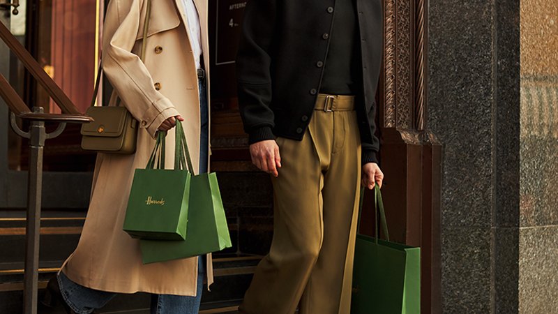 Two people in stylish outfits exit a building, each holding Harrods shopping bags.