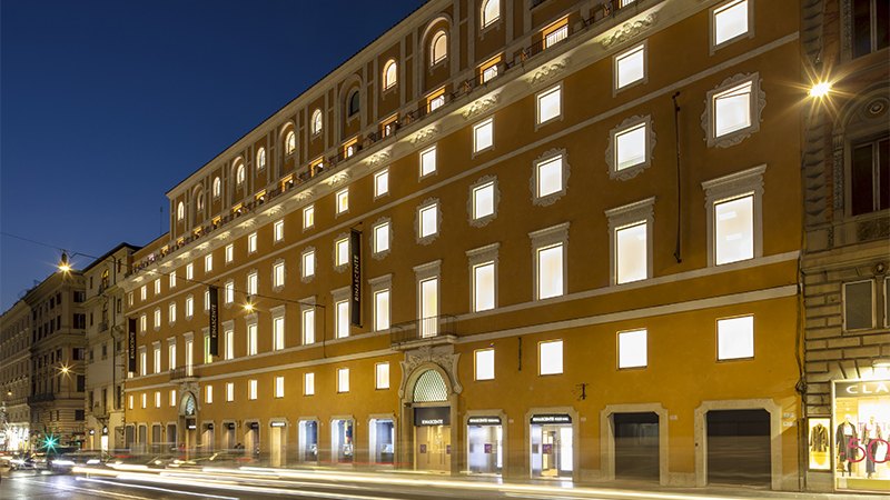 Illuminated facade of a historic building at night with arched windows and warm lights, on a bustling street with blurred car lights in Rome.