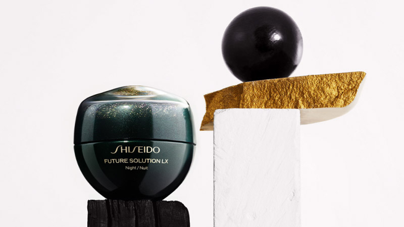 A Shiseido Future Solution LX jar sits elegantly on a black pedestal beside a textured white and gold block with a black orb on top.
