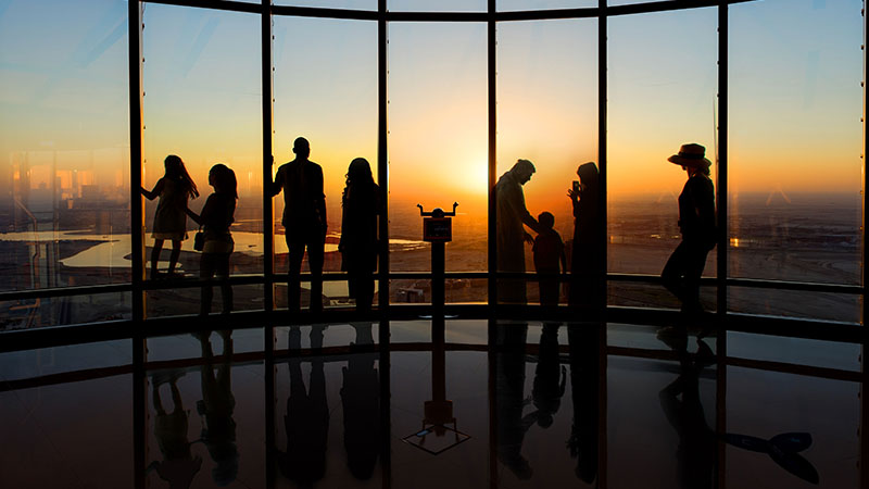 Silhouettes of people stand by large observation windows at sunset, overlooking a cityscape and water below.