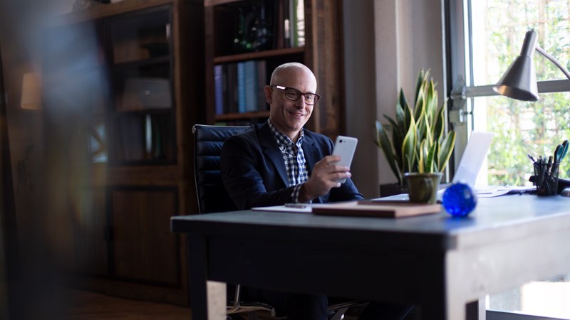 Man seated at desk, looking at his phone and smiling.